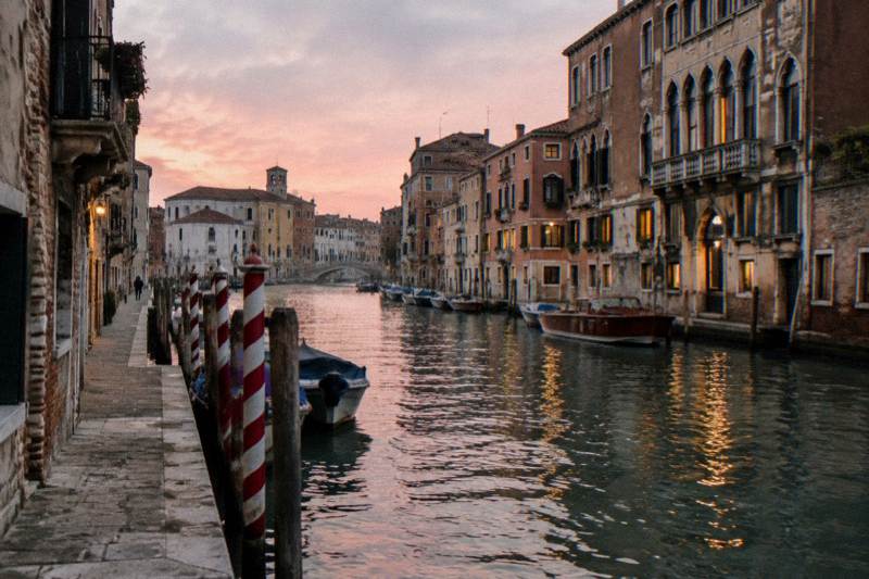 Soft pink dusk along the Cannaregio Canal beside Campo San Geremia in Venice, Italy, a wide waterway with moored wooden boats, tall palazzi with warm lit windows reflected in the water, candy-striped mooring poles in the foreground, one tiny distant figure on the fondamenta.