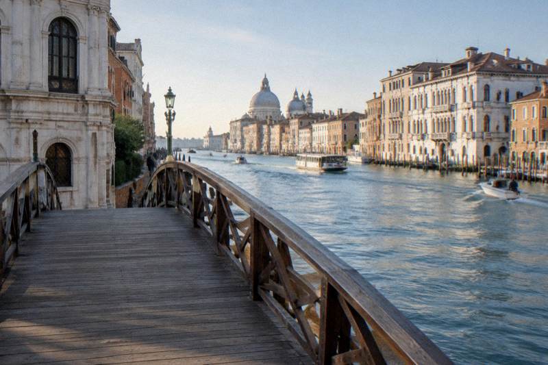 Clear morning at the foot of the arched wooden footbridge across the Grand Canal beside the Gallerie dell'Accademia in Dorsoduro, Venice, Italy, grand pale palazzi lining the far bank, boats moving on the water, the bridge's timber deck empty, nobody close to camera.