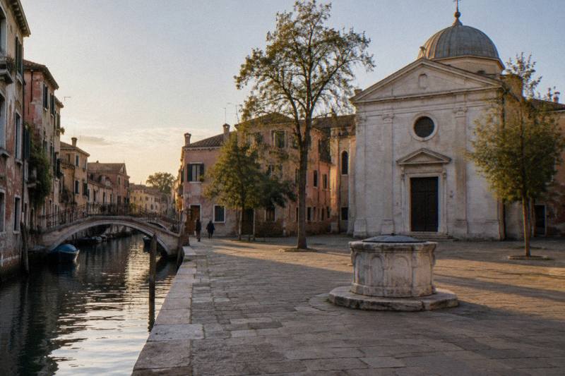 Late afternoon at Campo della Maddalena in Cannaregio, Venice, Italy, a small square with a plain neoclassical stone church and shallow dome on one side, a stone wellhead, pastel plaster houses and a few slender trees, long golden light, empty paving.