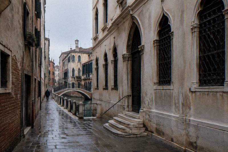 Early morning in the narrow stone-paved calle beside the Teatro Goldoni in San Marco, Venice, Italy, tall plastered facades with Gothic ogival windows and iron-barred shutters, the street only a few metres wide under a thin strip of grey sky, damp stones after rain.