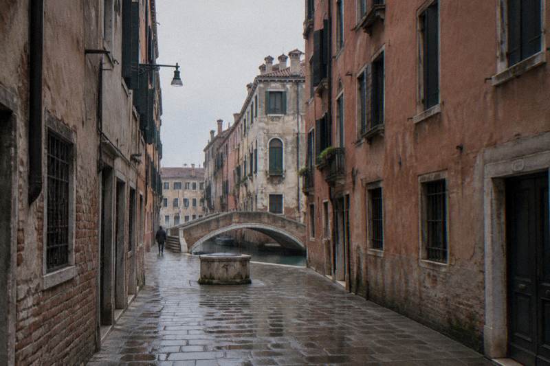 Overcast afternoon in a narrow calle near Campo San Geremia in Cannaregio, Venice, Italy, very tall flat-faced plastered tenements with rows of small shuttered windows pressing in on both sides, a small stone footbridge over a quiet side canal at the end of the lane, damp paving.
