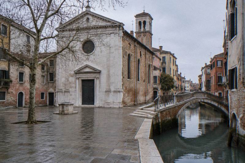 Grey soft morning at Campo de la Fava in Castello, Venice, Italy, a small square with a pale Istrian-stone church facade and brick side walls, a stone footbridge over a narrow canal at the corner, a bare plane tree, wet paving, nobody in frame.