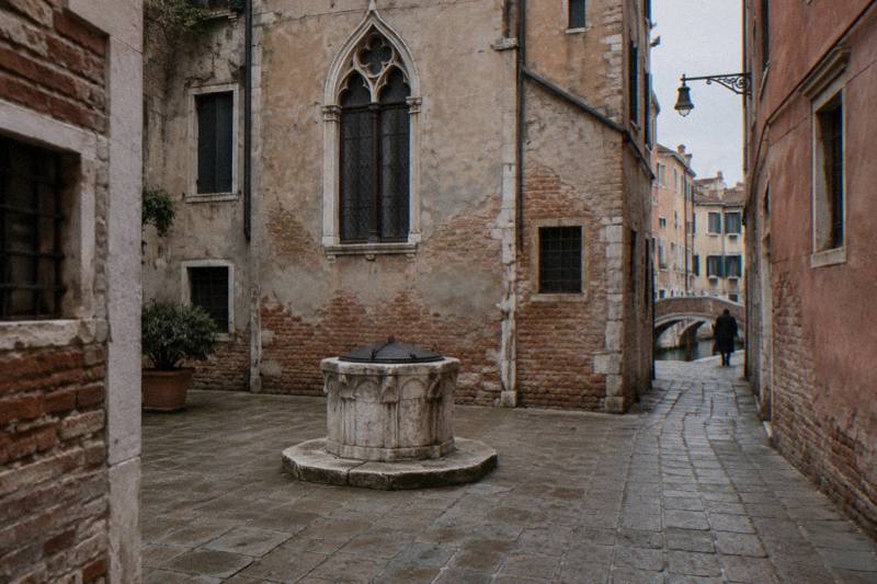 Soft overcast light in tiny Campiello de la Chiesa in San Marco, Venice, Italy, an enclosed little square of brick and plaster walls with a Gothic arched window, a worn stone wellhead at its centre, a narrow alley opening off one corner, a distant figure from behind.