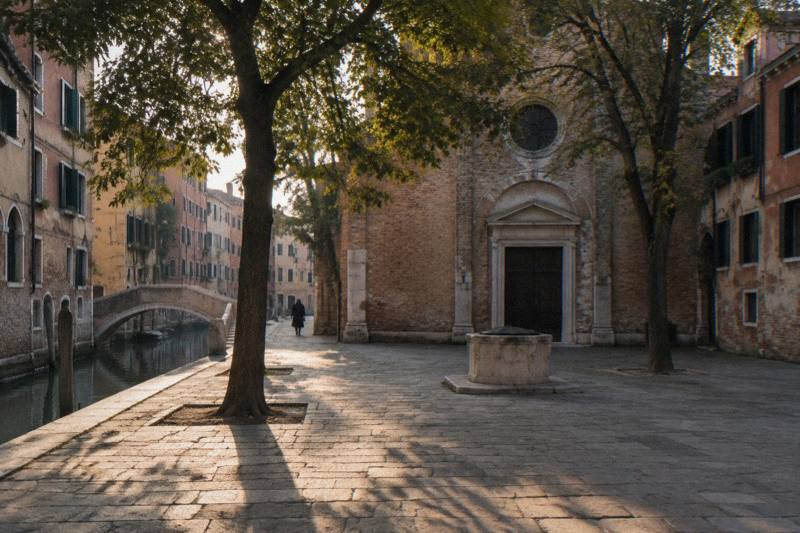 Late afternoon at Campo Sant'Agnese in Dorsoduro, Venice, Italy, a quiet shaded square with a few trees and a modest brick church front, faded plaster houses around it, warm sun slanting through the branches onto the paving, a lone distant figure walking away.