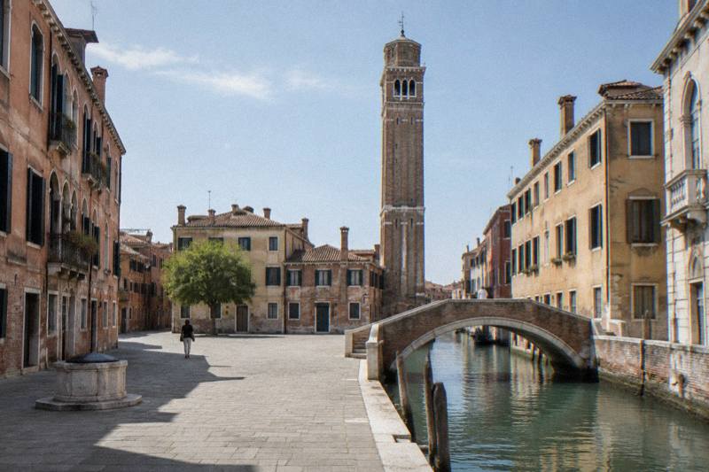Bright midday at Campo Santi Apostoli in Cannaregio, Venice, Italy, a tall slender brick bell tower rising above a wide square, a stone footbridge over the canal beside it, pale terracotta and ochre houses, a few distant figures crossing the paving.