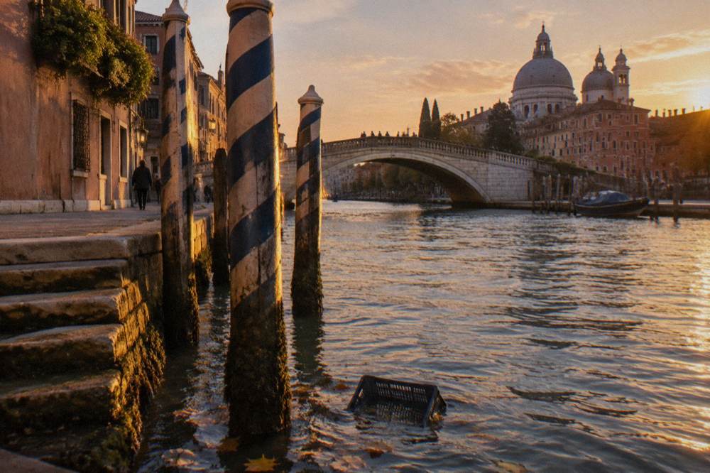 A Venice canal at water level with mooring posts and a stone bridge