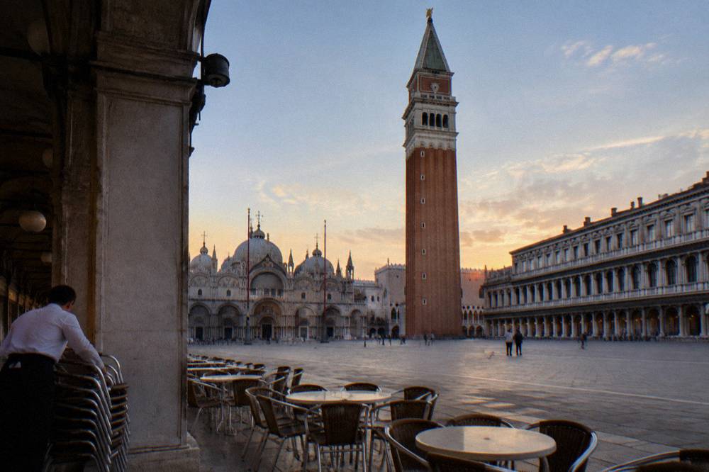 A wide stone square with arcaded buildings and a tall brick bell tower under an evening sky