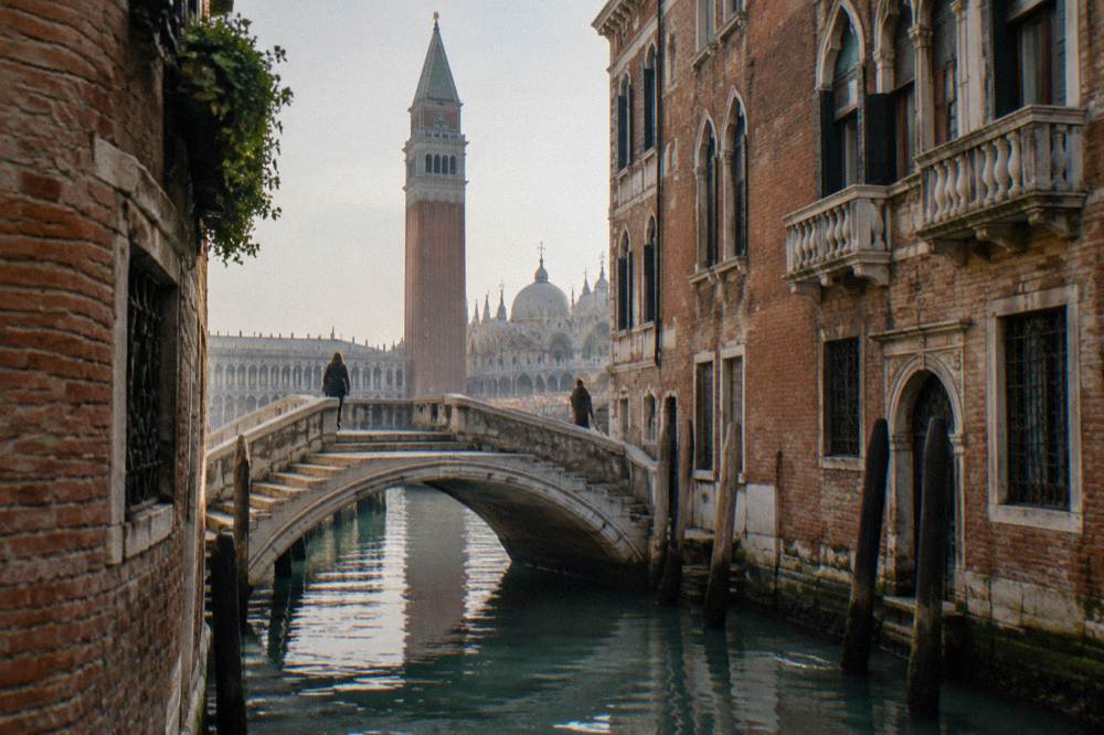 A narrow canal between old brick buildings with a small stone bridge and mooring posts