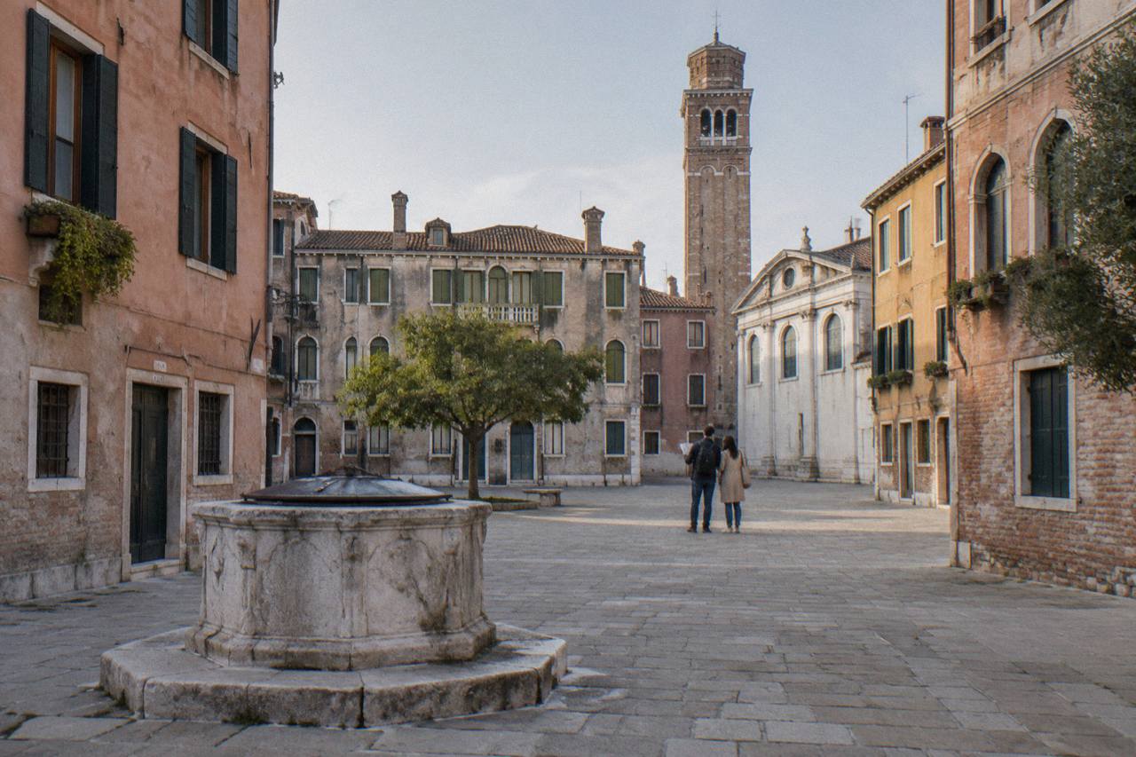 A worn stone well head in a quiet Venice campo with pastel shuttered facades and a brick bell tower beyond