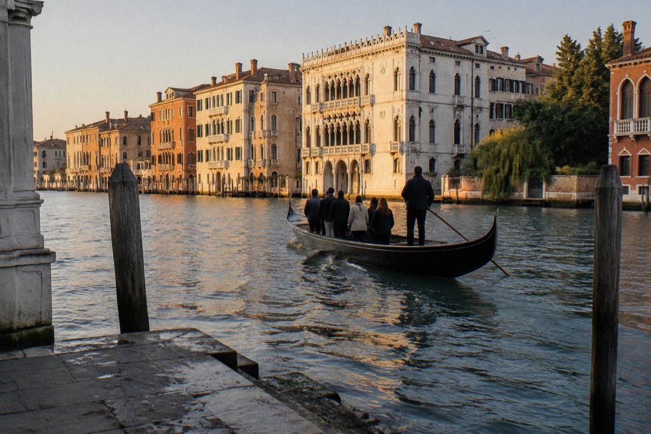 A black gondola carrying standing passengers across the Grand Canal in Venice at golden hour, seen from a stone landing
