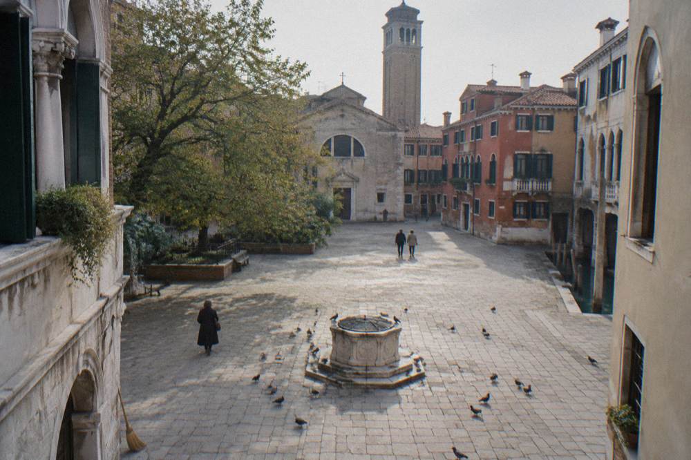 A sunlit Venice campo with a well head in the centre and pigeons on the paving