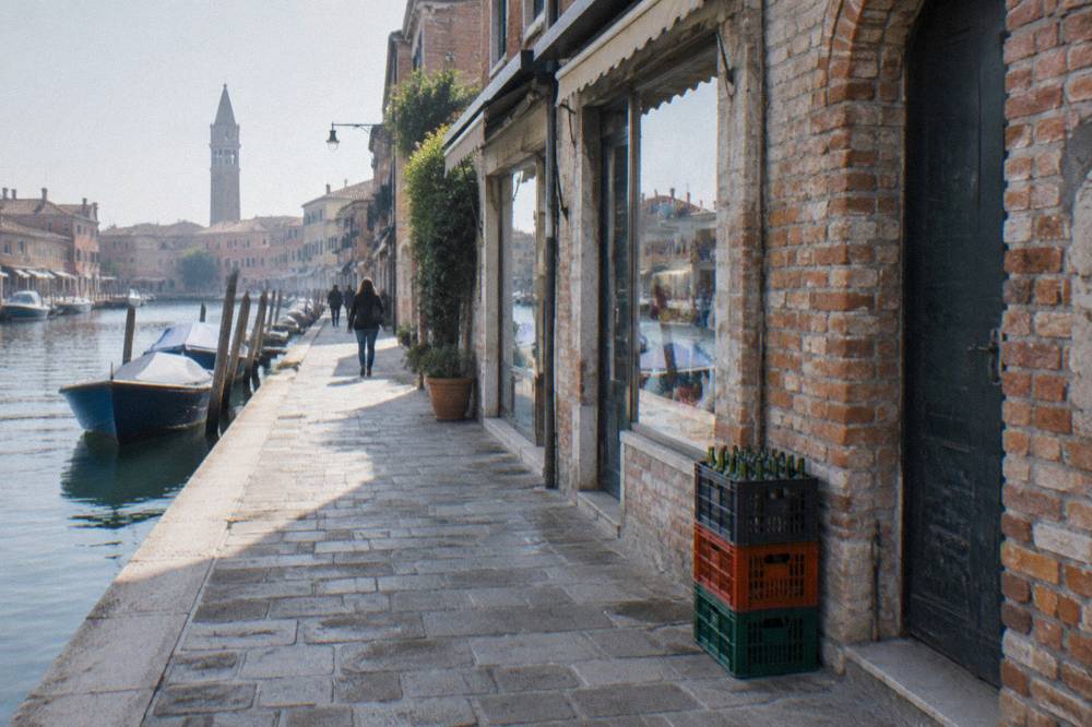 A waterfront walkway in Murano lined with old brick furnace buildings and shop fronts along a quiet canal