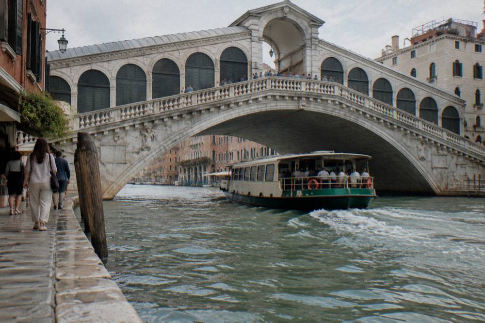 A vaporetto waterbus passing under the Rialto Bridge with passengers on the open deck