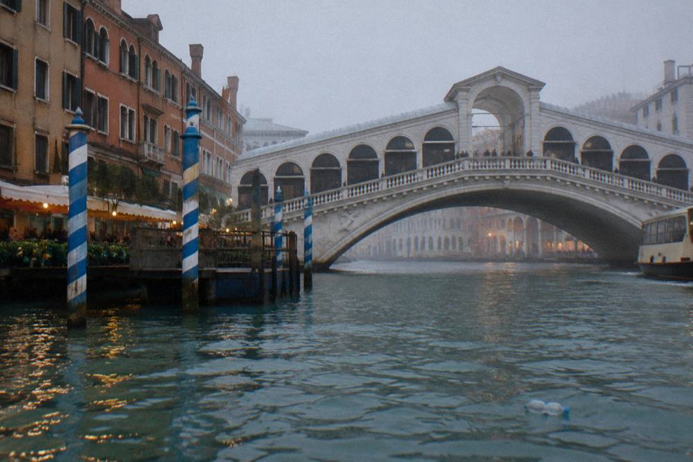 The Rialto Bridge seen from a quiet stretch of the Grand Canal with striped mooring poles in the water