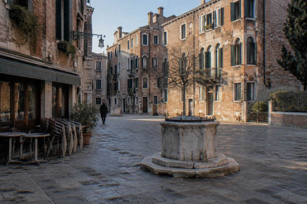 A Venice campo with a well head in the centre and cafe tables under arcade shadows
